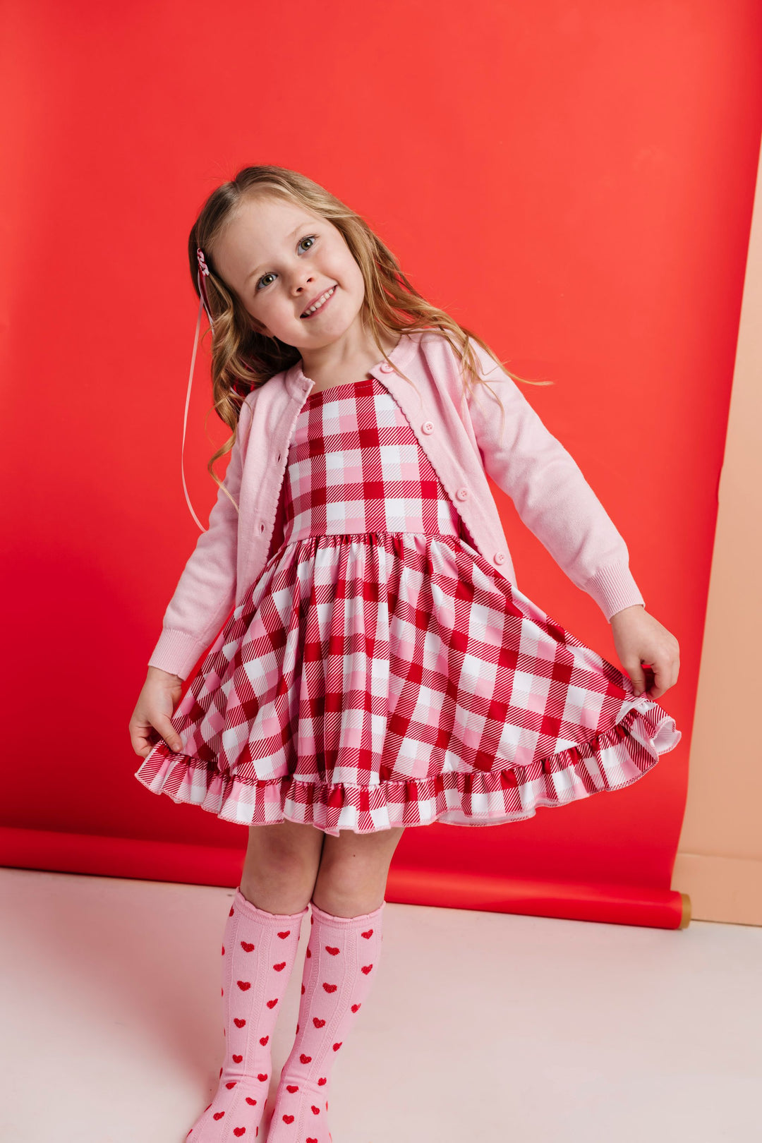 girl posing in red, white and pink plaid twirl dress paired with light pink cotton knit cardigan and matching pink knee highs with red hearts