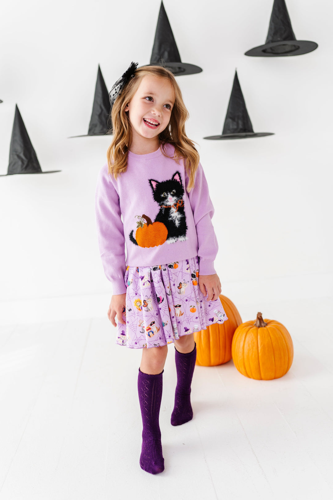 young girl surrounded by pumpkins and floating witch hats wearing lavender twirl dress with kittens dressed in Halloween costumes paired with matching purple fluffy kitten and pumpkin sweater, purple pointelle knee socks and sheer black star bow