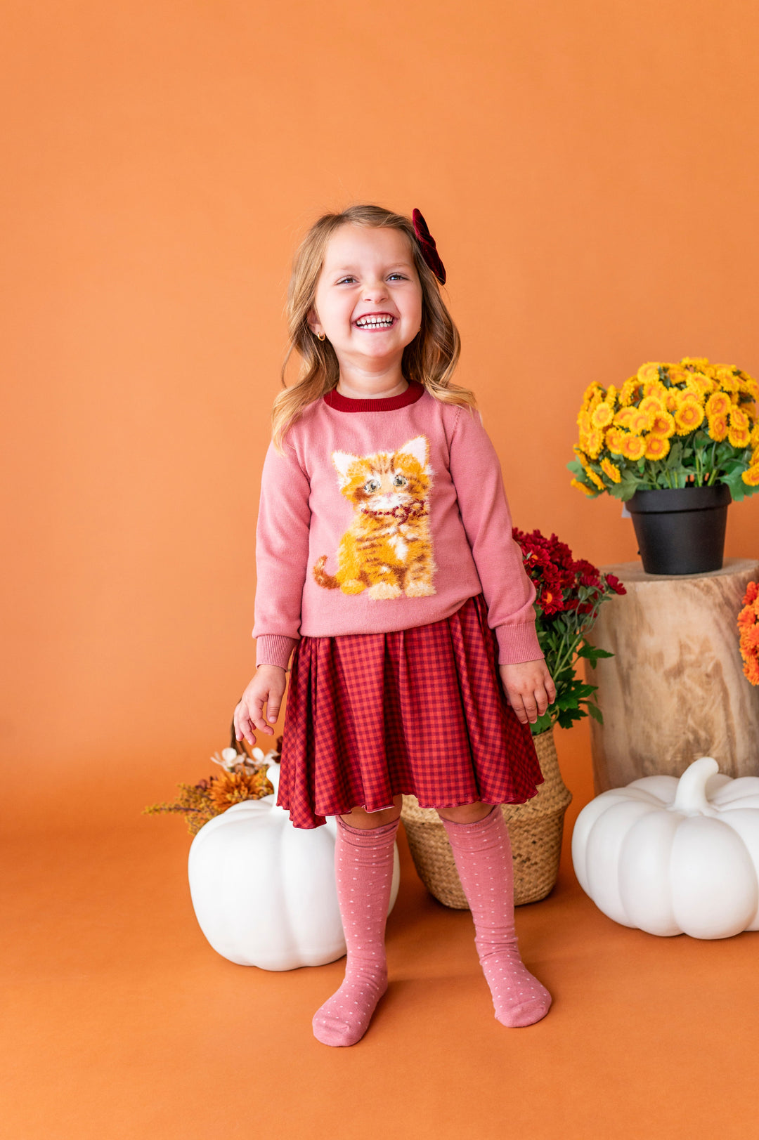 young girl in front of fall flowers wearing pink kitten sweater paired with rust gingham fall twirl dress and matching pink dot knee socks