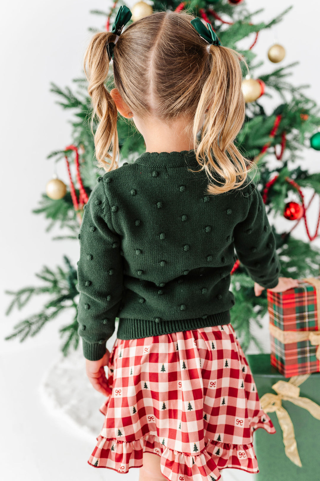 A young girl with blonde pigtails stands with her back to the camera, wearing a dark green textured knit sweater and a red and white gingham skirt with a small Christmas tree print. She is posed in front of a decorated Christmas tree with ornaments and a wrapped gift box.