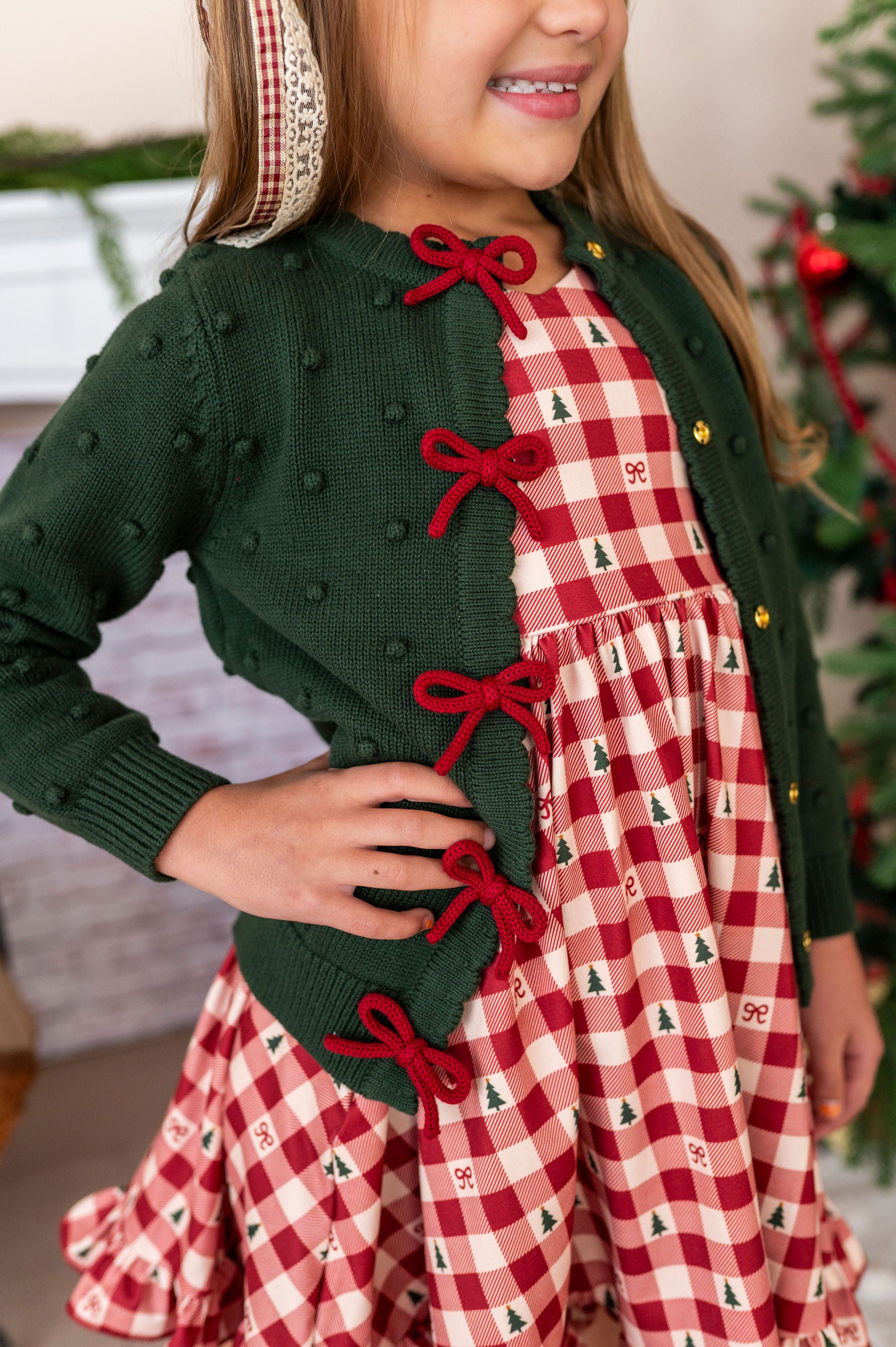 little girl with hand on hip wearing a dark green knit cardigan with crimson bows down the placket over the snap buttons and a checkered christmas tree and bow dress
