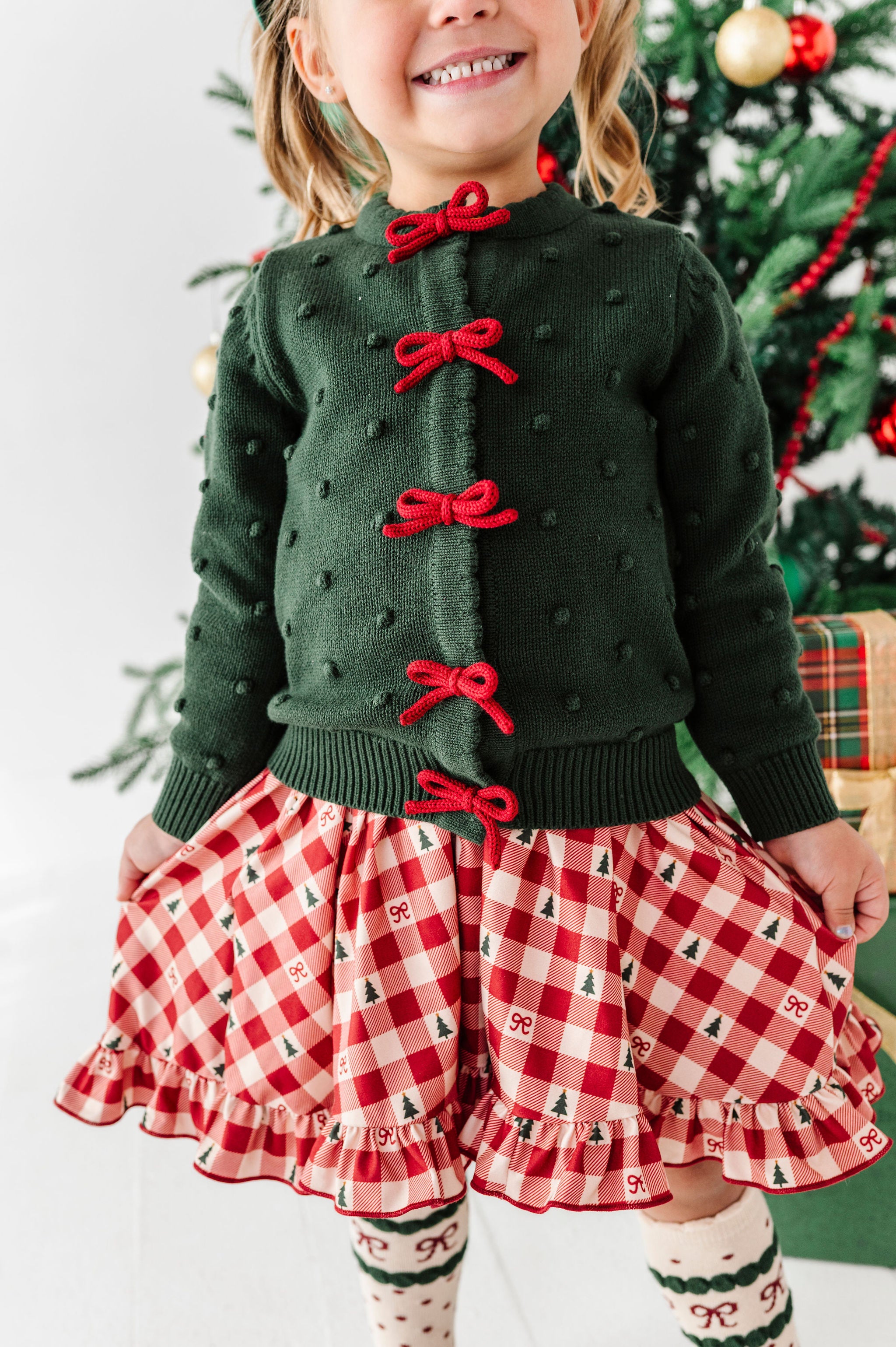 A young girl wearing a festive Christmas outfit, featuring a dark green knit cardigan with red bow details and a red-and-white gingham skirt with a small tree pattern, standing in front of a decorated Christmas tree.