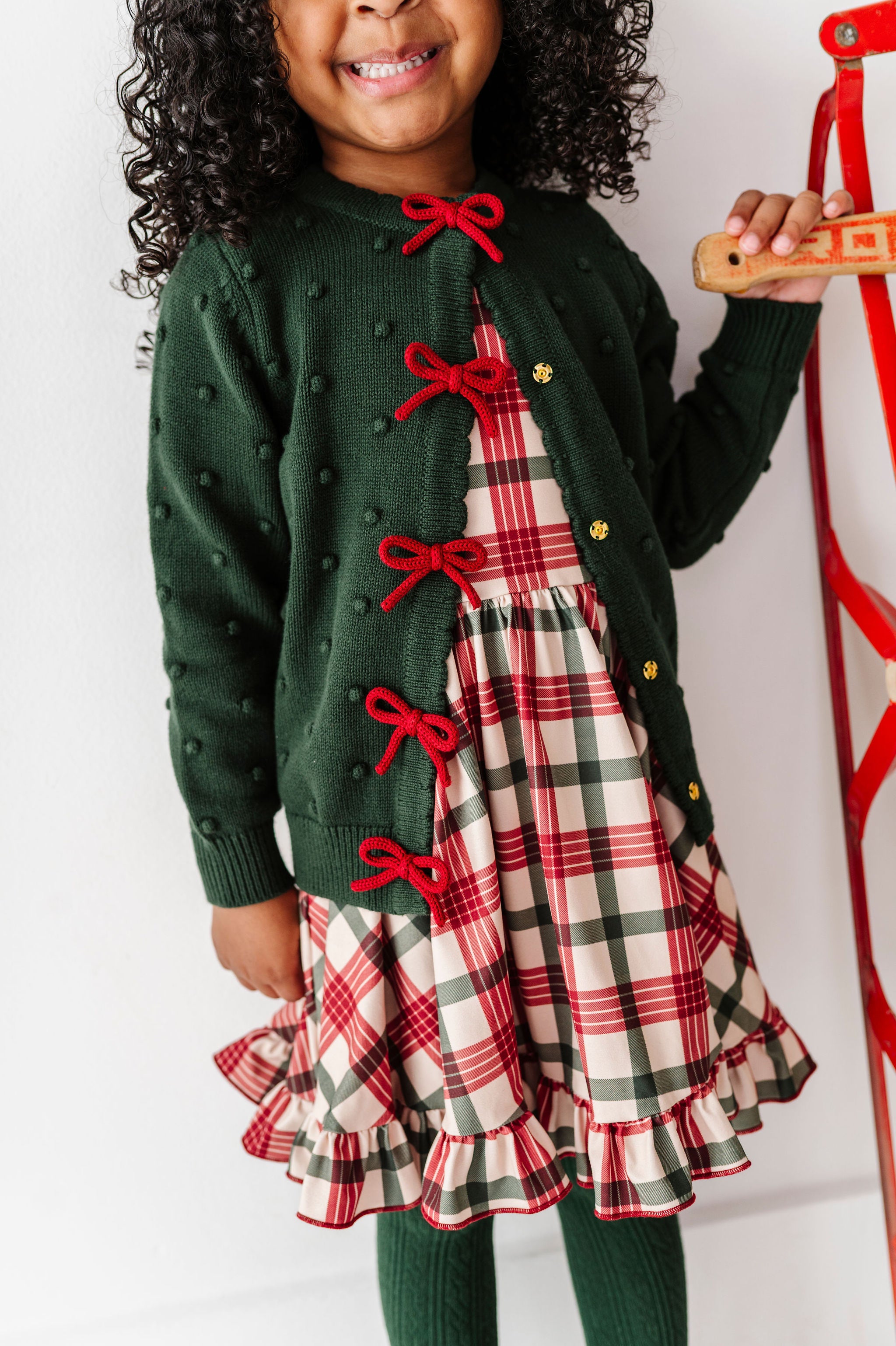 A young child wearing a festive holiday outfit consisting of a dark green knit cardigan with red bow details, a red, green, and white plaid dress, and matching green tights, standing next to a red sled