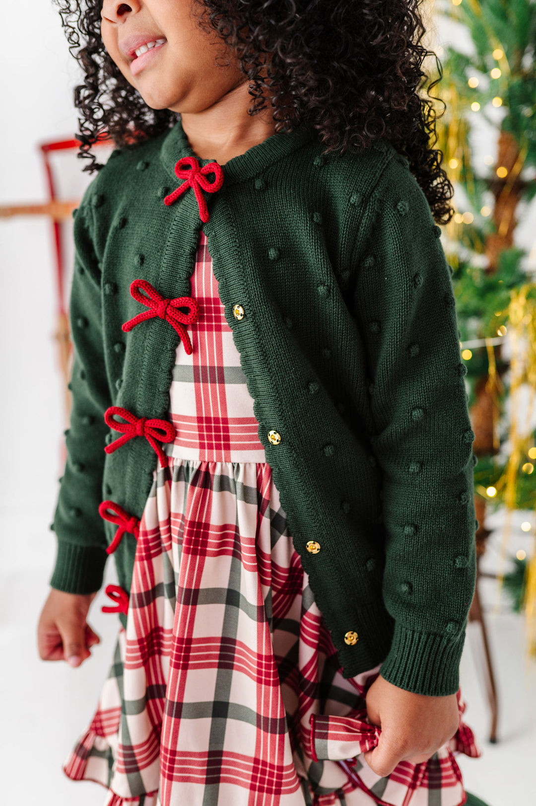 A child wearing a dark green knit cardigan with red bow details and gold buttons, layered over a red, white, and black plaid dress, in a festive holiday setting.
