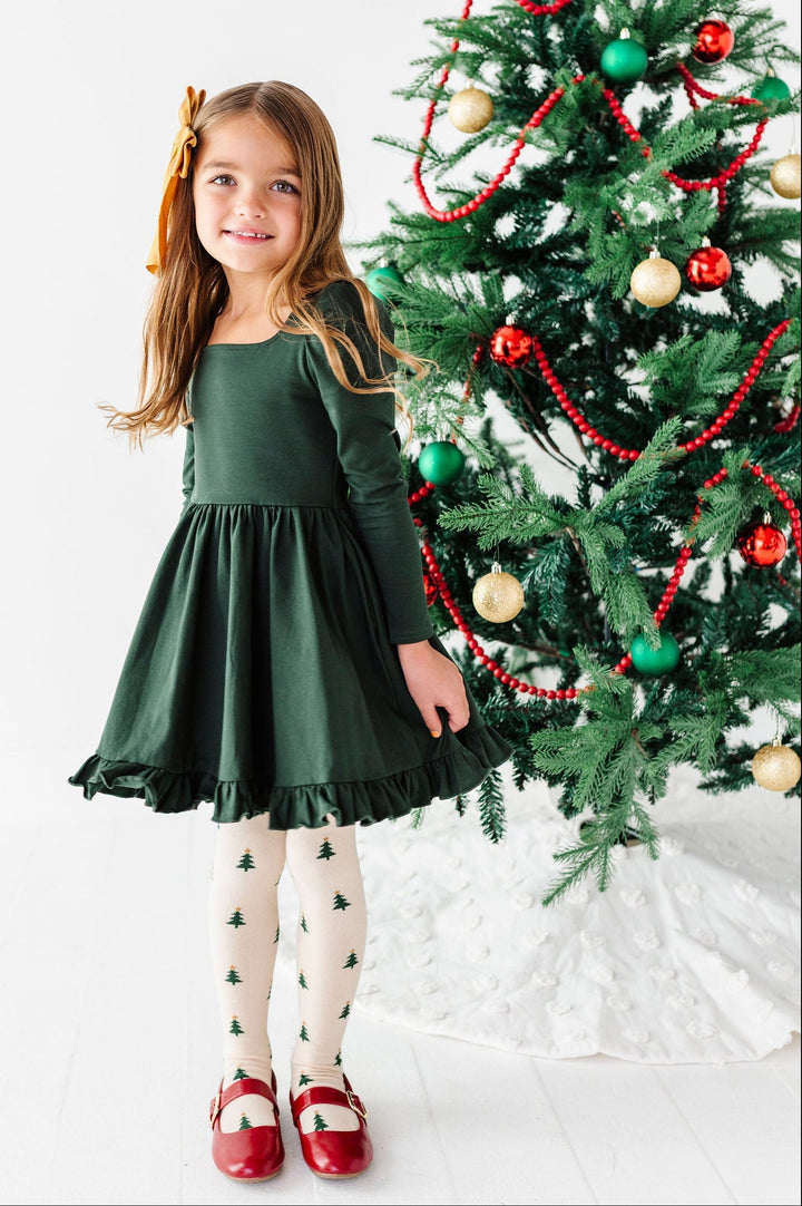 A young girl stands next to a decorated Christmas tree wearing a dark green long-sleeve dress, white tights with small green tree prints, and red shoes.