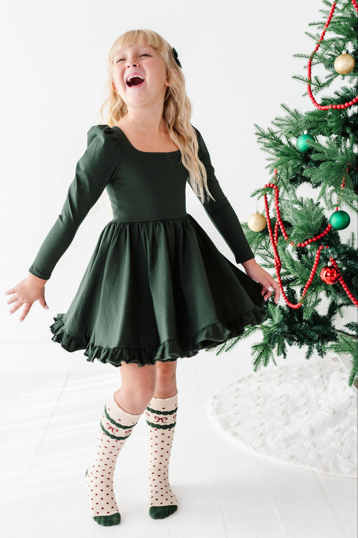A young girl with blonde hair wearing a dark green long-sleeve dress and patterned socks, laughing joyfully next to a decorated Christmas tree in a bright white room.