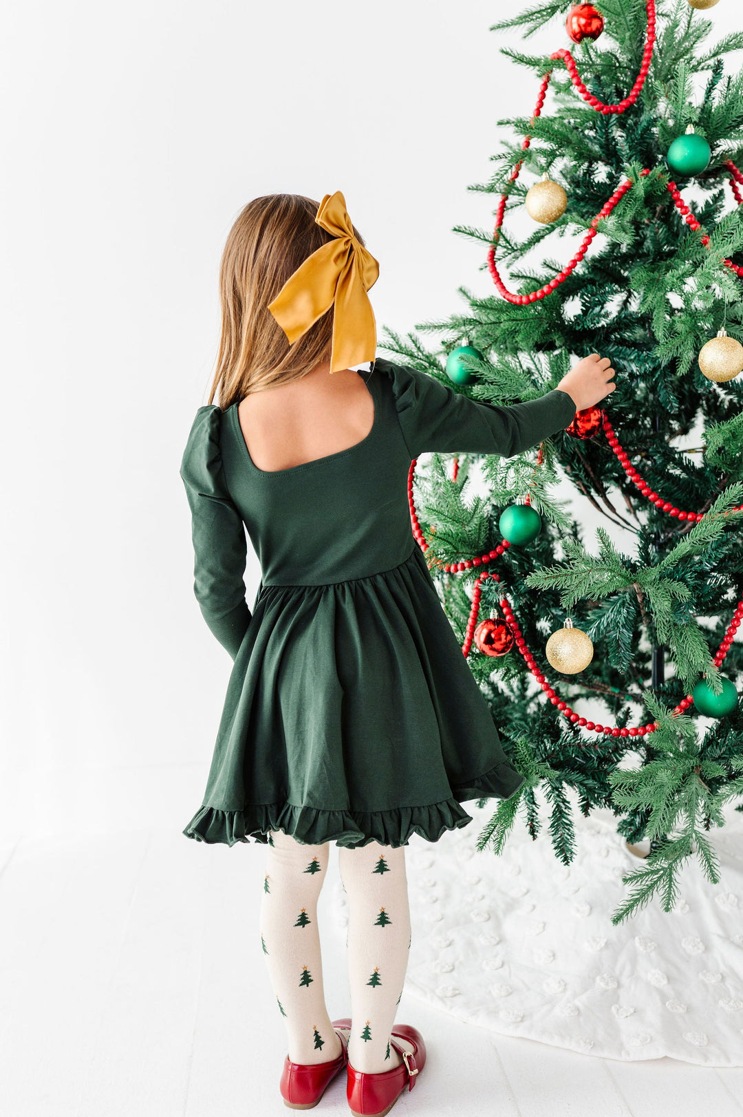 A young girl viewed from behind wearing a dark green long-sleeve dress, white tights with a small green tree pattern, and red shoes, standing next to a decorated Christmas tree and wearing a large yellow bow in her hair.