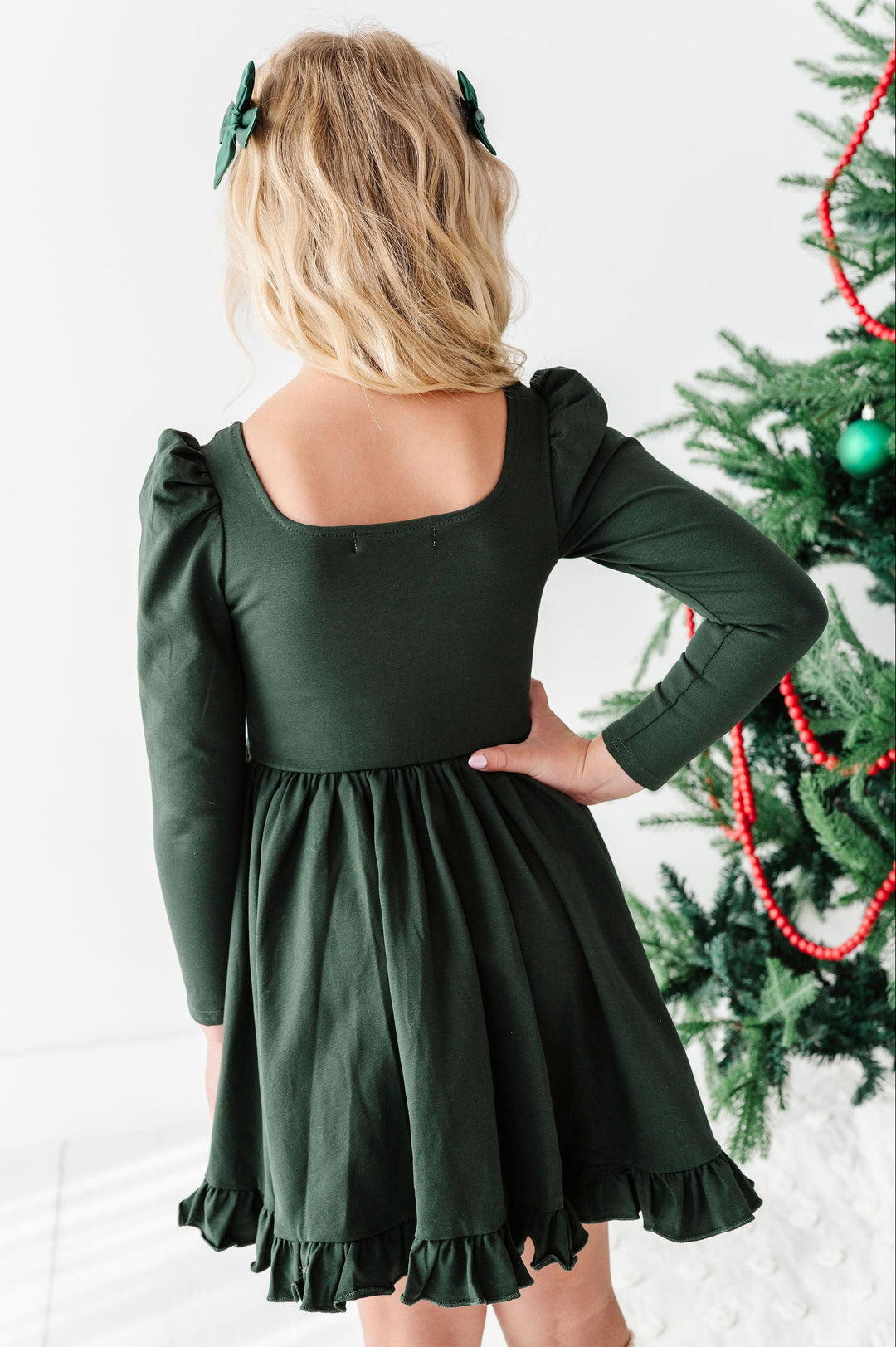 A young girl with blonde hair wearing a dark forest green long-sleeve dress, shown from behind. She is standing next to a decorated Christmas tree in a bright, white indoor setting.