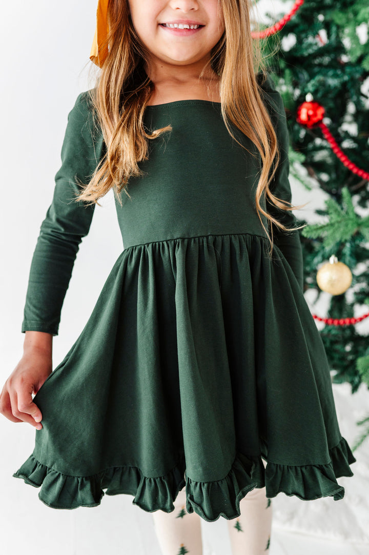 A young girl wearing a dark forest green long-sleeve dress with a ruffled hem, standing in front of a decorated Christmas tree.