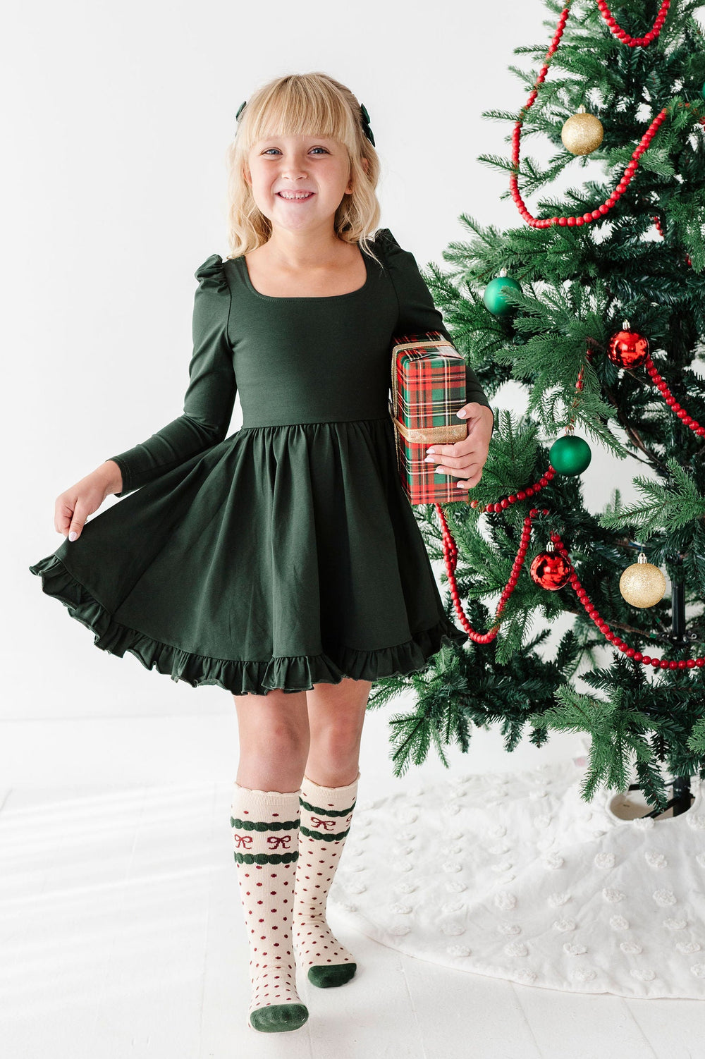 A young girl with blonde hair stands next to a decorated Christmas tree, wearing a dark green long-sleeve dress, patterned knee-high socks, and holding a wrapped gift box.