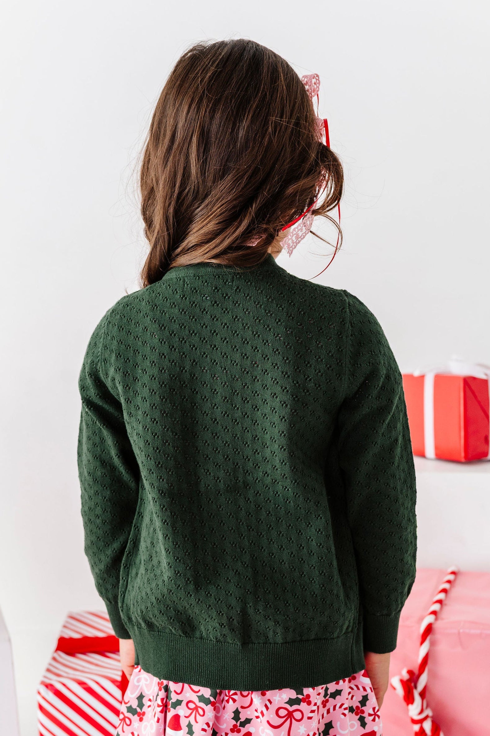 A young girl viewed from behind wearing a dark green textured knit sweater, a pink and white floral skirt, and white leggings with a small green pattern. She is standing in a festive setting surrounded by red and white wrapped gift boxes.