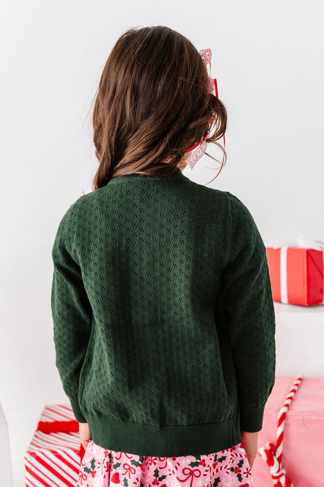 A young girl viewed from behind wearing a dark green textured knit sweater, a pink and white floral skirt, and white leggings with a small green pattern. She is standing in a festive setting surrounded by red and white wrapped gift boxes.