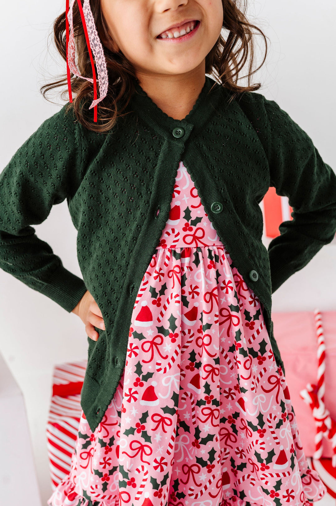 A young girl wearing a dark green knit cardigan over a pink Christmas print dress, standing in a festive indoor setting with holiday decorations.