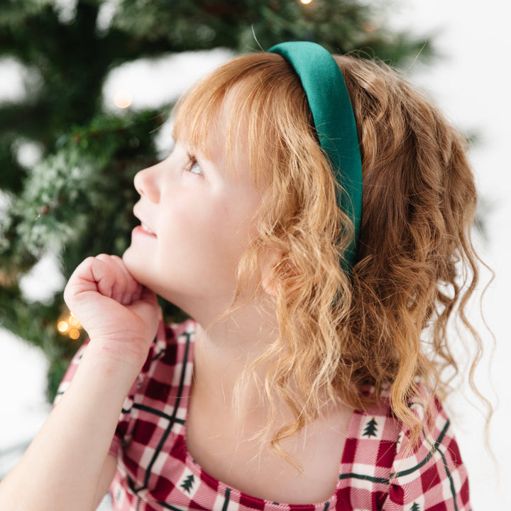 little girl sitting by Christmas tree in green satin headband and red gingham Christmas dress