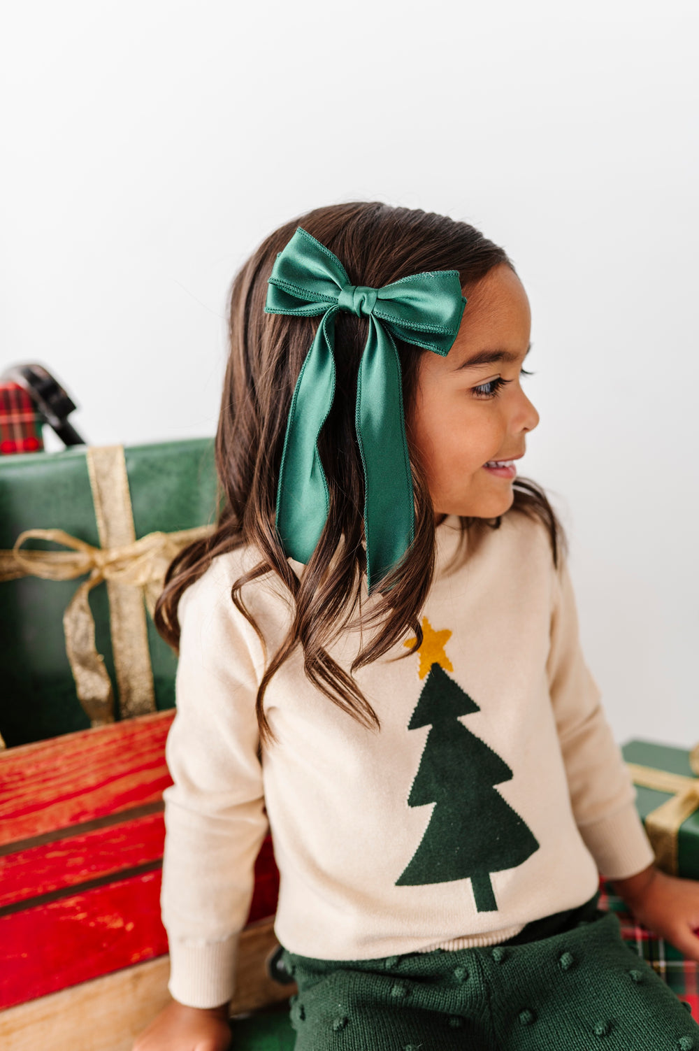 A young girl wearing a dark green satin bow headband and a cream knit sweater with a green felt Christmas tree, sitting next to wrapped Christmas presents.