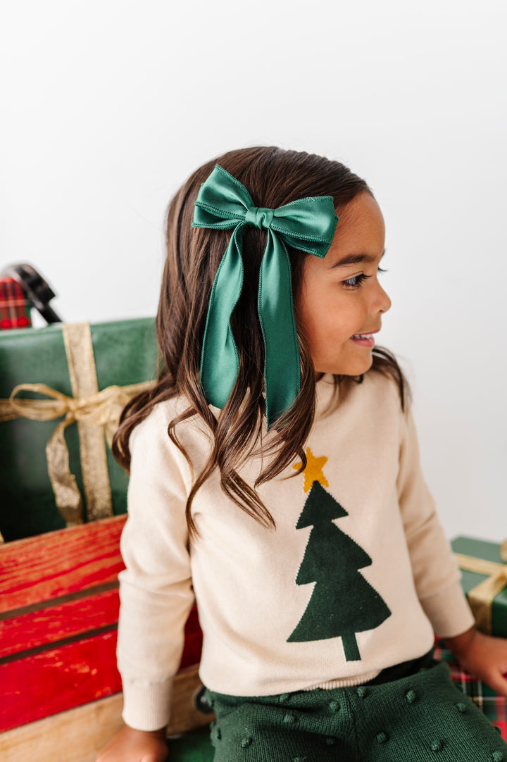 A young girl wearing a dark green satin bow headband and a cream knit sweater with a green felt Christmas tree, sitting next to wrapped Christmas presents.