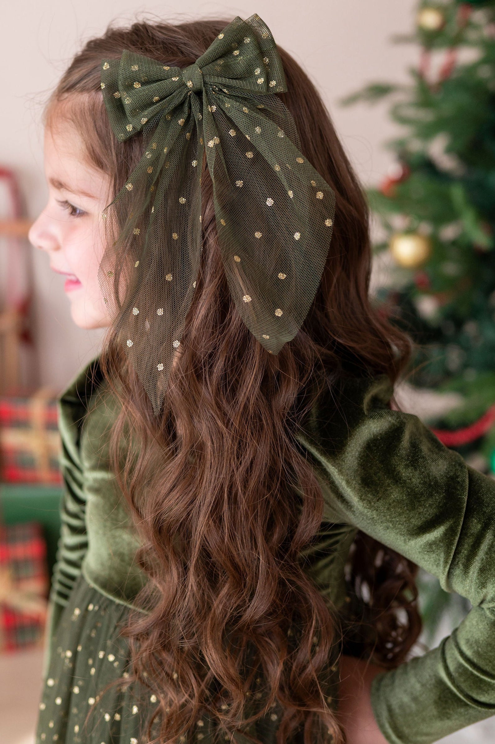 little girl wearing forest velvet and tulle xmas dress with a matching forest and gold tulle hair bow set against a blurred holiday background