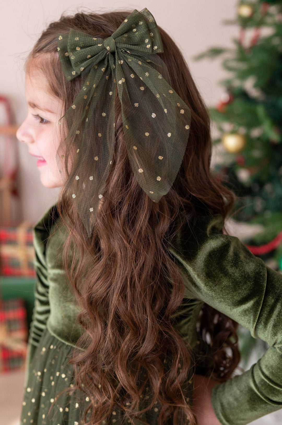 little girl wearing forest velvet and tulle xmas dress with a matching forest and gold tulle hair bow set against a blurred holiday background