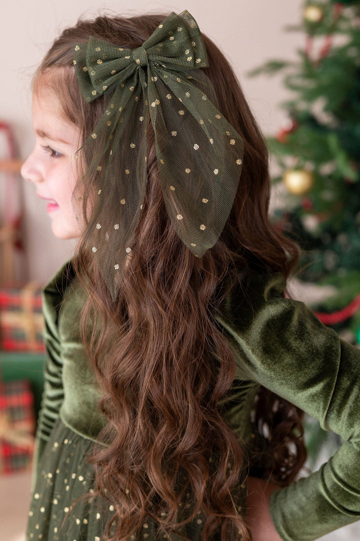 little girl wearing forest velvet and tulle xmas dress with a matching forest and gold tulle hair bow set against a blurred holiday background