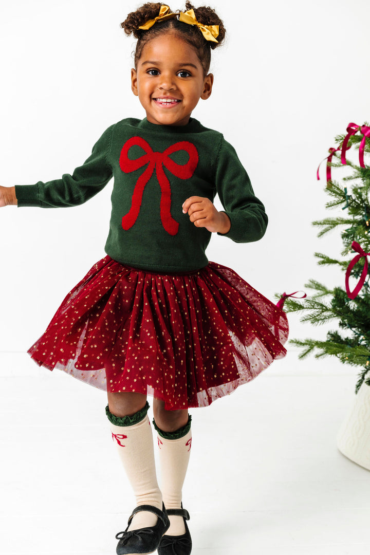 girl twirling in crimson and forest green Christmas bow sweater, matching crimson tutu skirt with gold sparkly dots, gold satin pigtail bows and bow knee highs with green lace trim