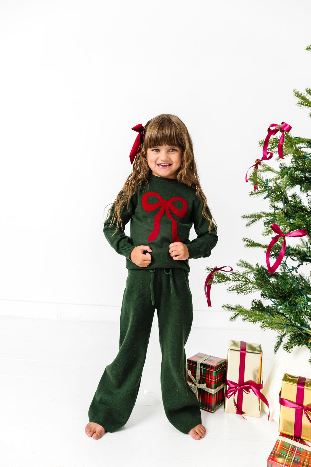 little girl standing by Christmas tree in green pullover sweater with red bow design, matching green sweater pants and red satin hair bow 