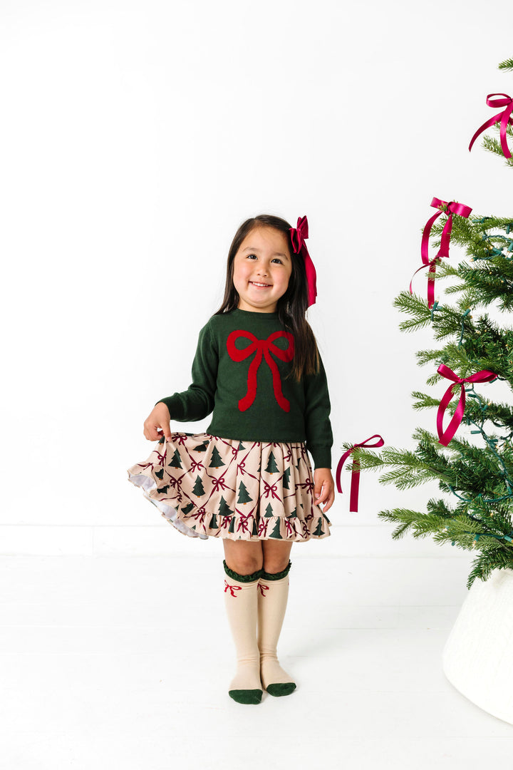 little girl by Christmas tree in forest green sweater with crimson bow design paired with matching tree and bow print twirl dress, red satin bow and knee highs with bow design