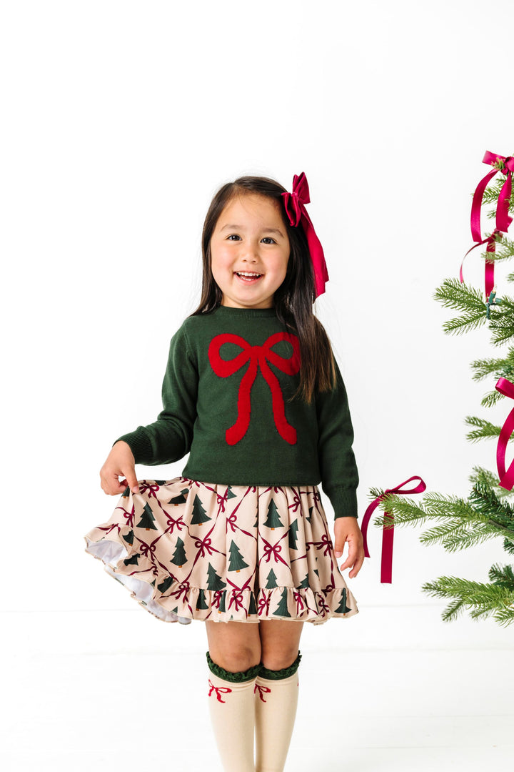 young girl by Christmas trees in forest green sweater with crimson bow design paired with matching bow sicks, cream tree and bow print twirl dress and crimson satin hair bow