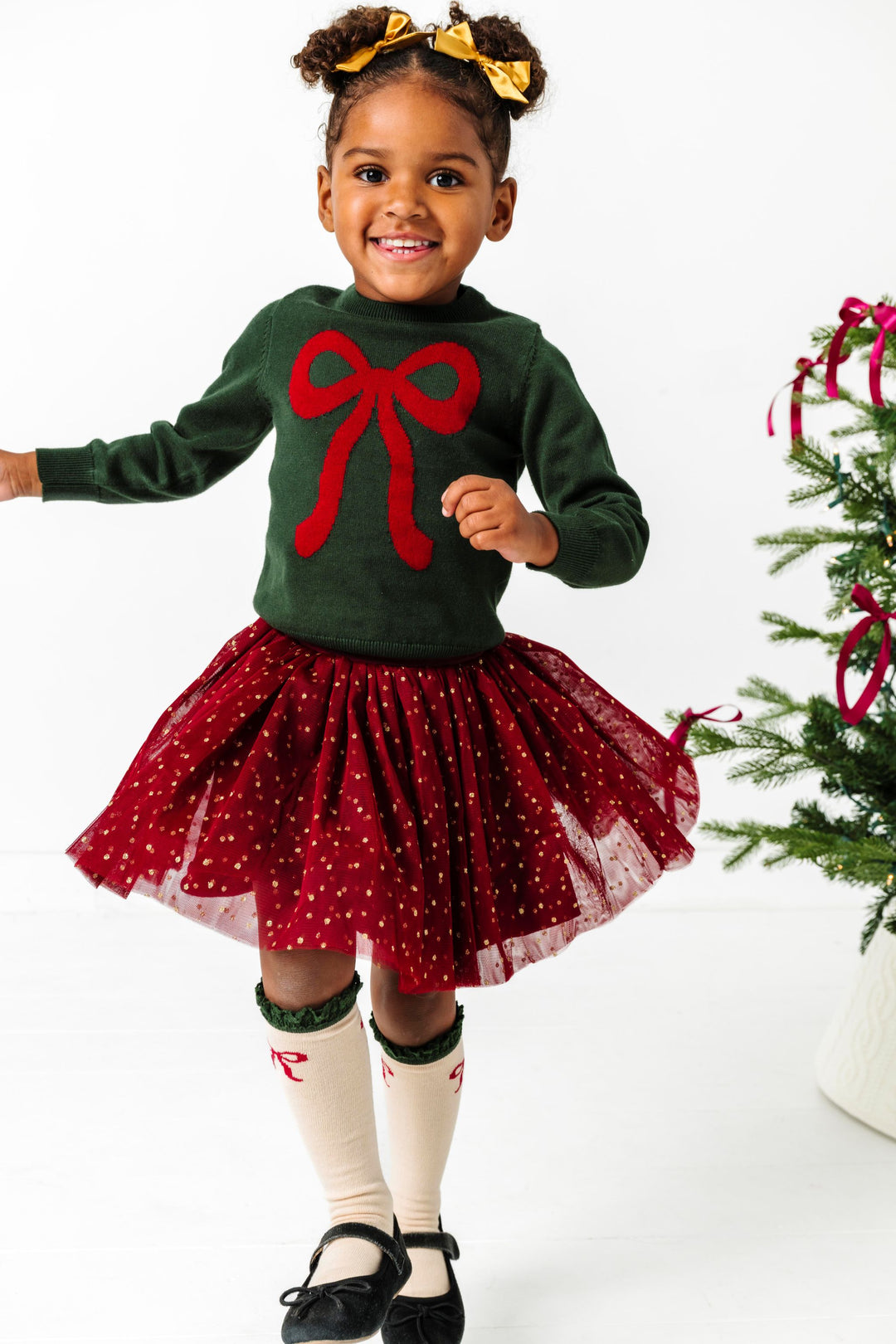 little girl by Christmas tree in forest green sweater with crimson bow design paired with crimson tulle sparkle skirt, gold satin pigtail bows and bow knee highs