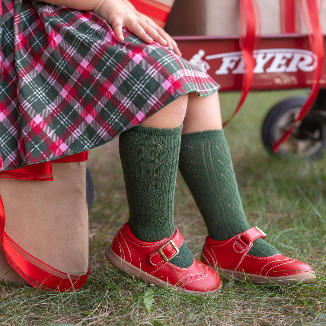 little girl sitting on Christmas gift wearing forest green open knit knee high socks and matching Christmas plaid skirt