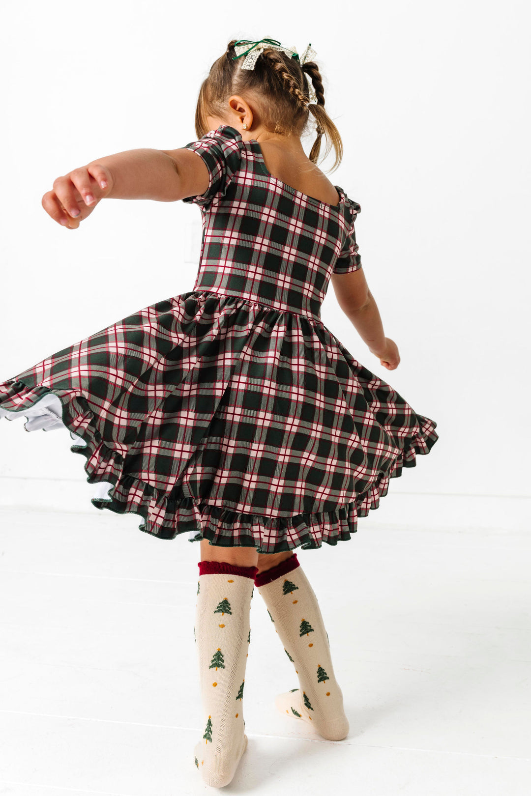 little girl twirling in forest green plaid short sleeve dress and cream socks with Christmas tree design and crimson scalloped trim