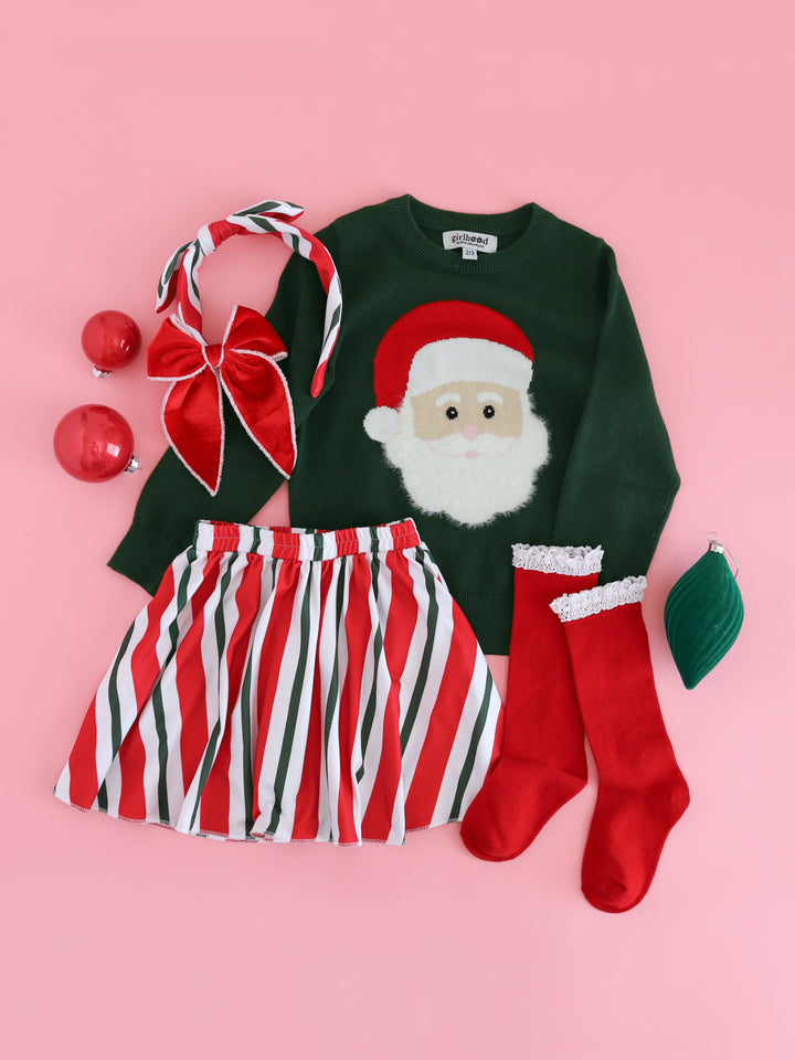 girls' Christmas outfit flat lay featuring forest green knit Santa sweater with fluffy beard, candy cane striped skirt and headband and red knee socks with white lace trim