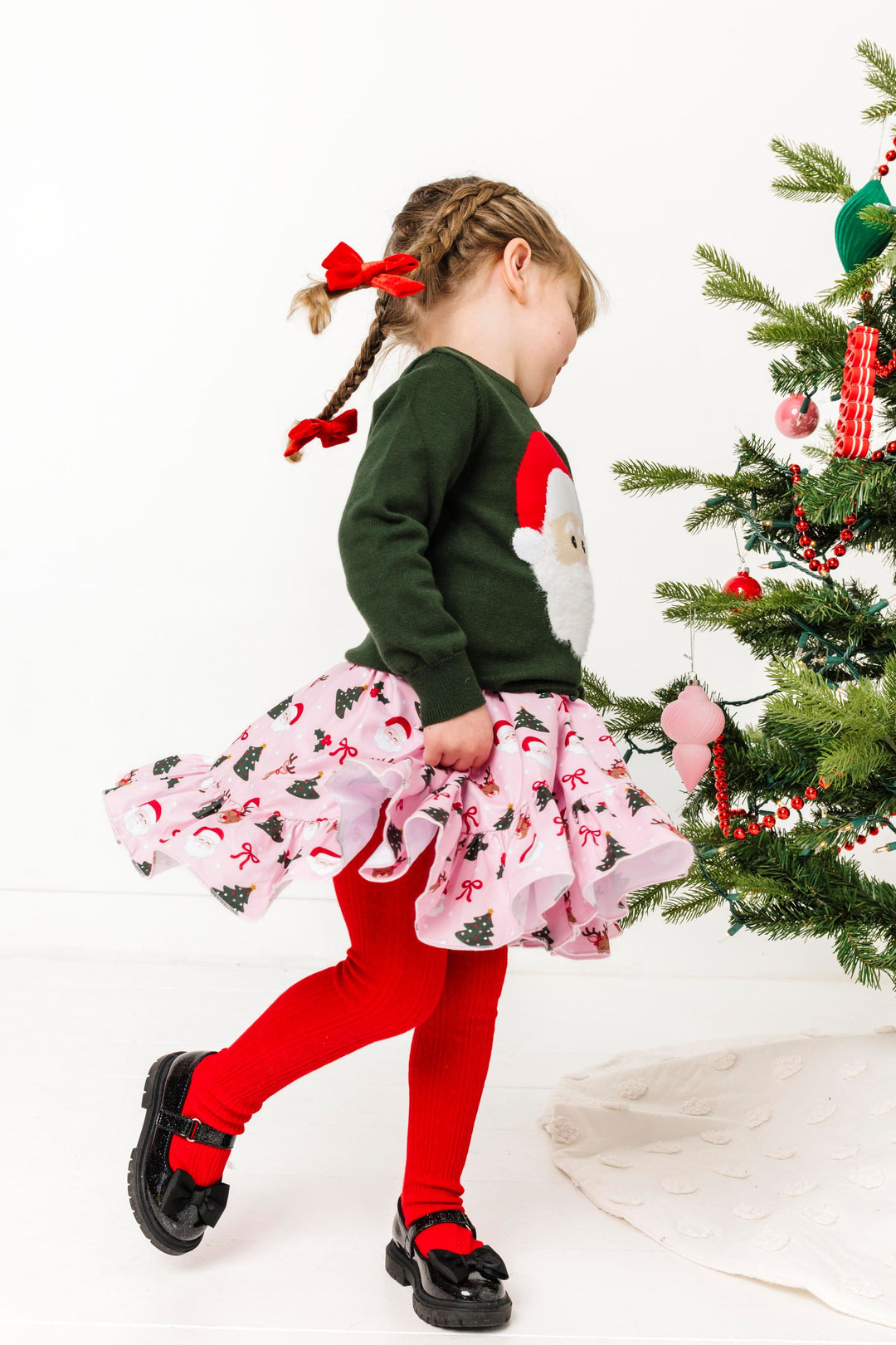 little girl in braids twirling by Christmas tree in forest green sweater with santa face, red cable knit tights and matching pink twirl dress with Santa and Rudolph print