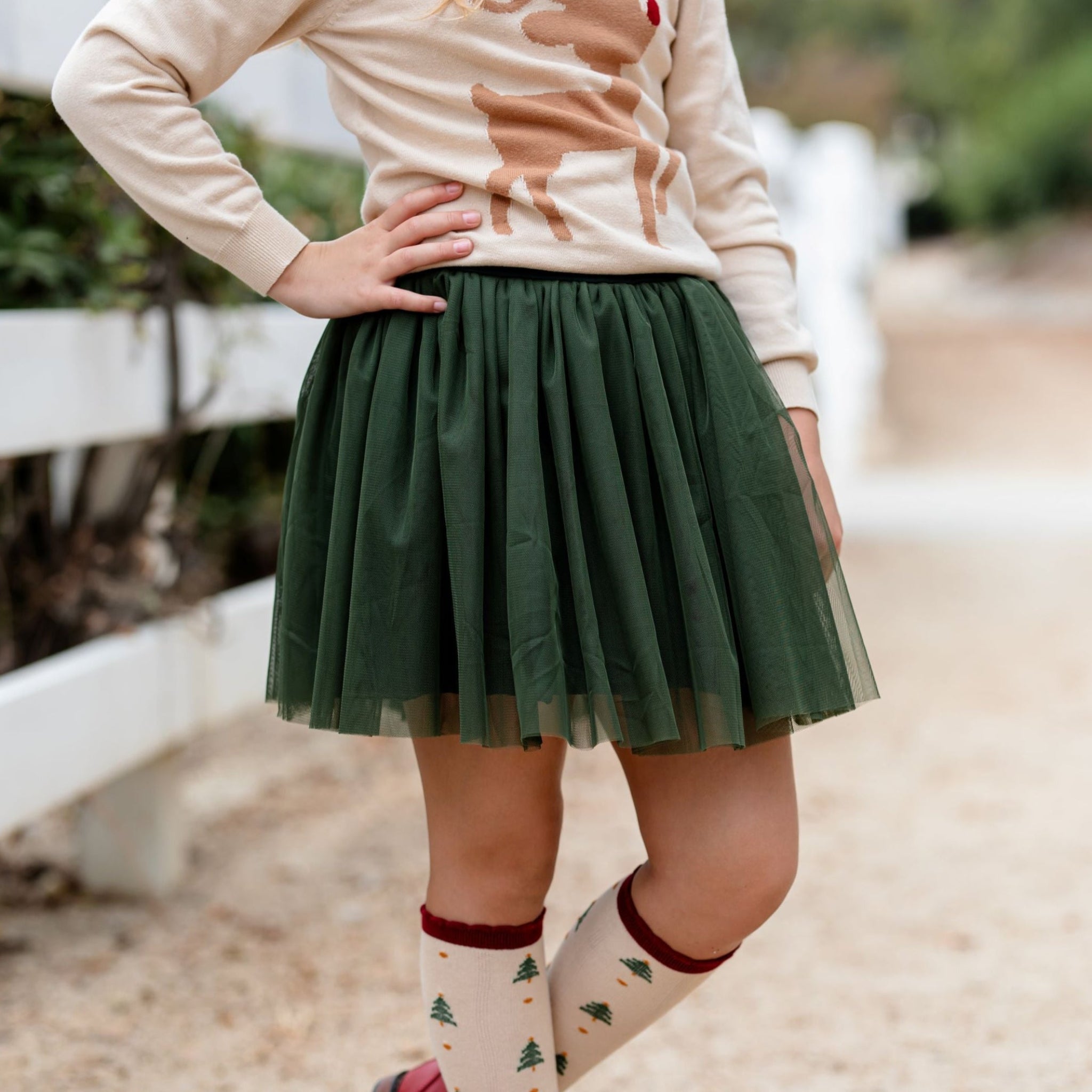 little girl in forest green tulle tutu skirt paired with cream reindeer sweater and matching Christmas tree knee high socks