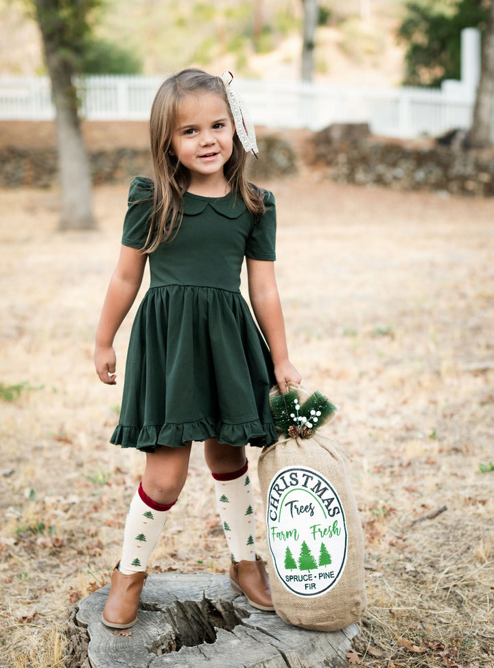 little girl at tree farm wearing forest green short sleeve collared twirl dress paired with matching Christmas tree print knee high socks