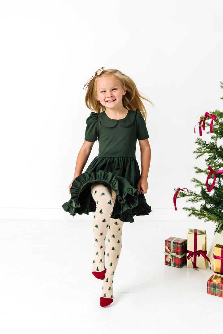 little girl skipping by Christmas tree in forest green short sleeve collared twirl dress paired with cream tights with green tree design and matching bow