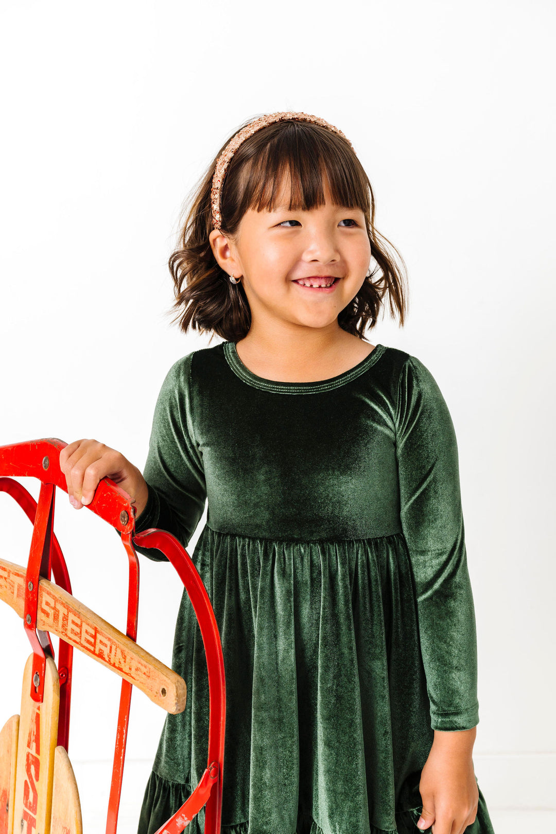 young girl with sled in forest green velvet long sleeve twirl dress and sparkly gold headband for Christmas