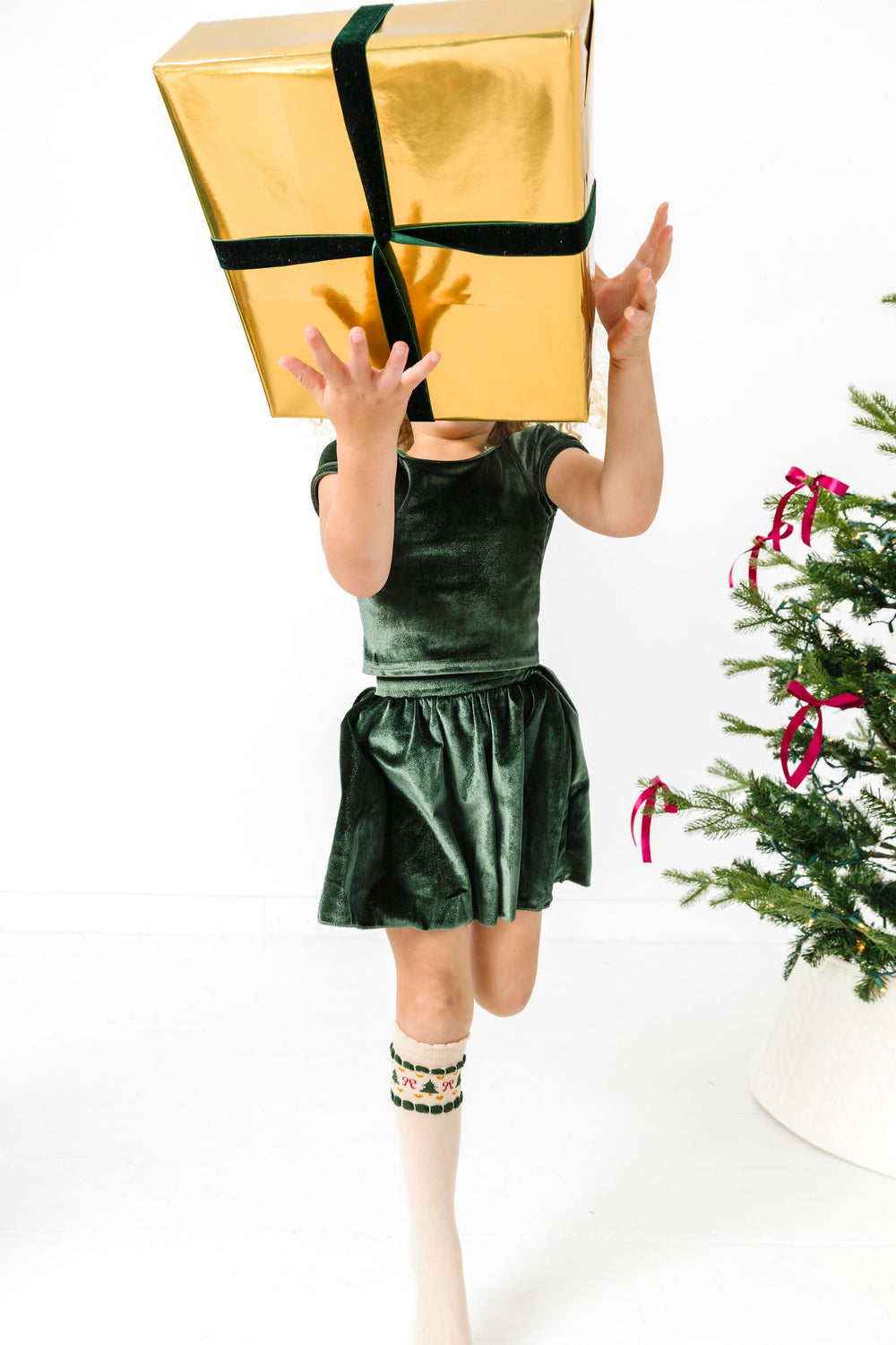 little girl jumping with gold gift box in forest green velvet top and skirt and matching Christmas themed fair isle knee socks