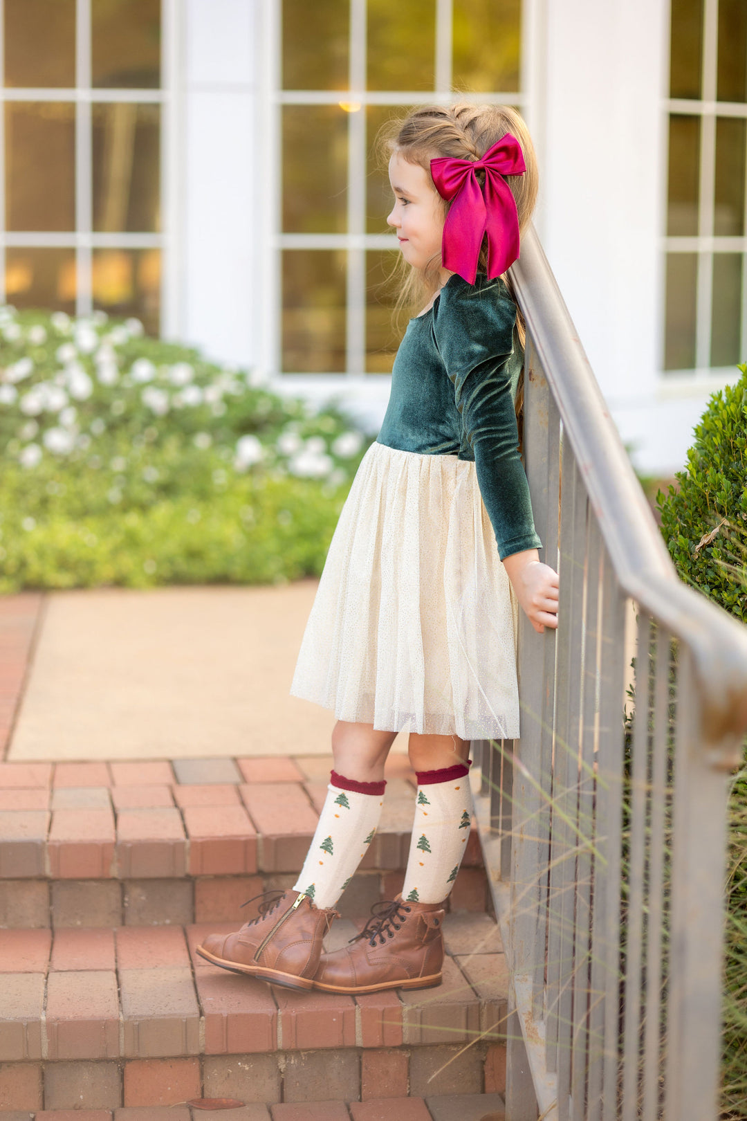 little girl wearing forest green Christmas dress with sparkly cream skirt, matching crimson satin hair bow and Christmas tree print knee high socks