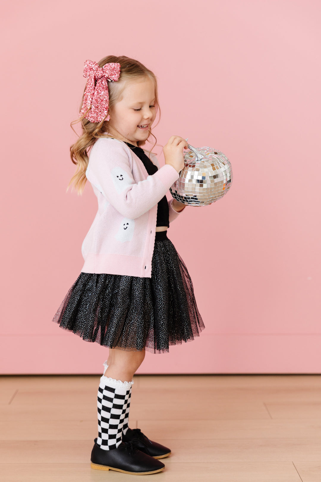 little girl holding mirrored silver pumpkin in pink knit v-neck cardigan with ghost print paired with black velvet top, black sparkle tutu skort, black checkered knee highs and pink sequin bow