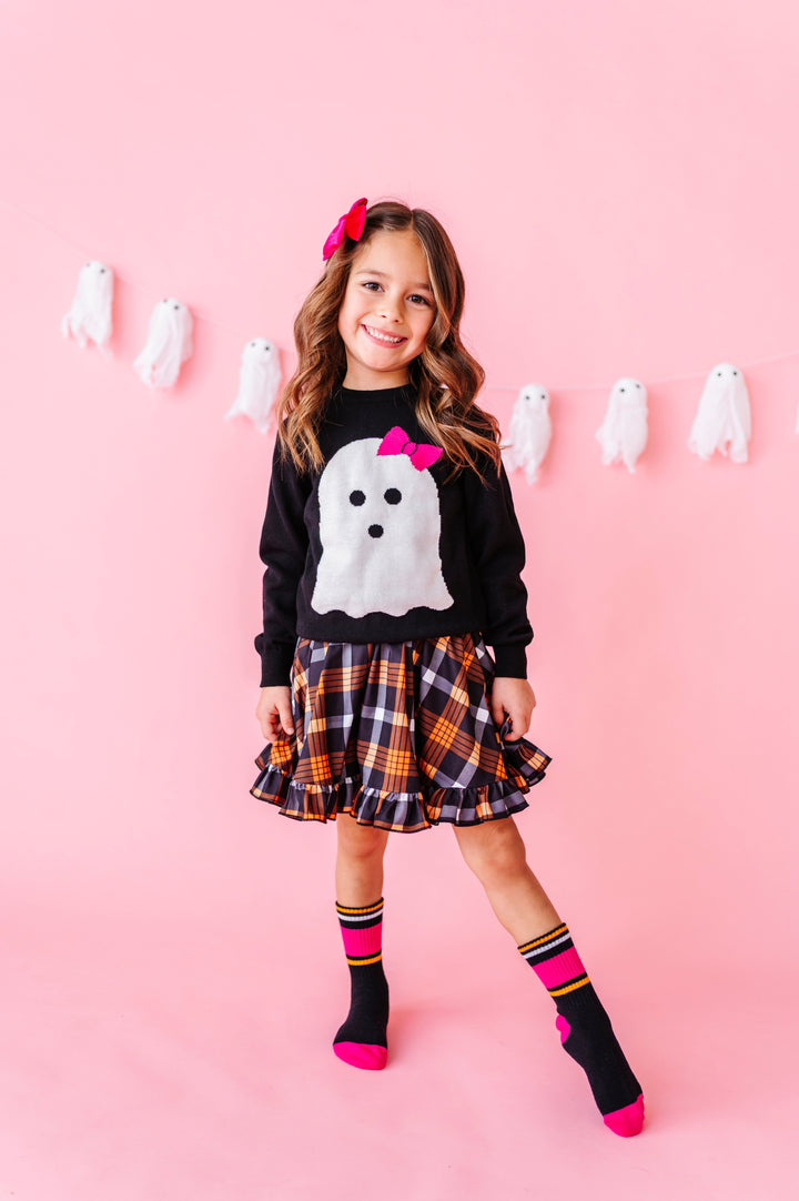 young girl in front of ghost garland wearing black cotton knit pullover sweater with white ghost wearing hot pink bow and paired with orange and black plaid dress and striped midi socks