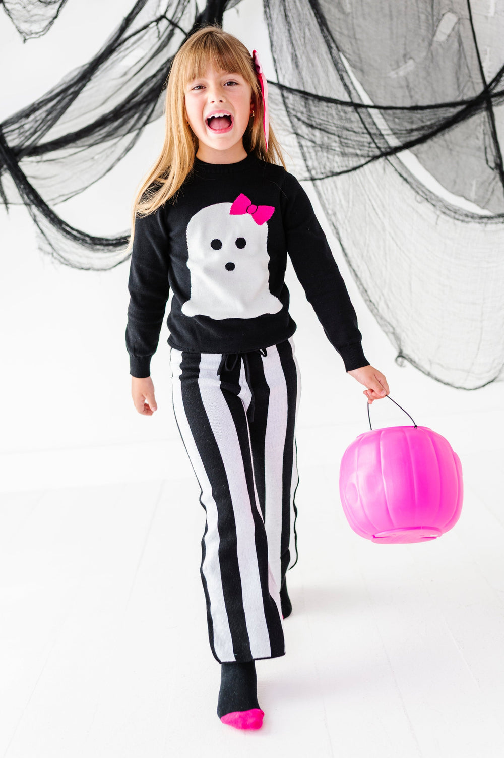 young girl holding pink pumpkin bucket in black and white striped sweater pants paired with white ghost design on black cotton knit sweater
