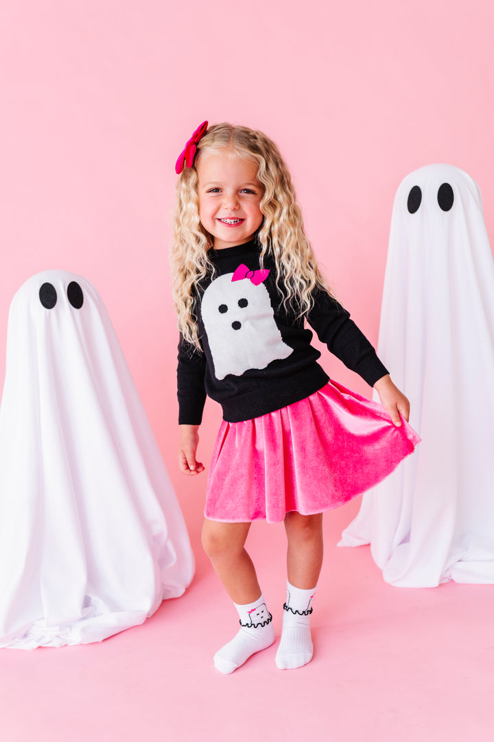 young girl standing beside sheet ghost decorations wearing black cotton knit sweater with white ghost wearing pink bow design paired with matching ghost midi socks, pink velvet skort and hair bow