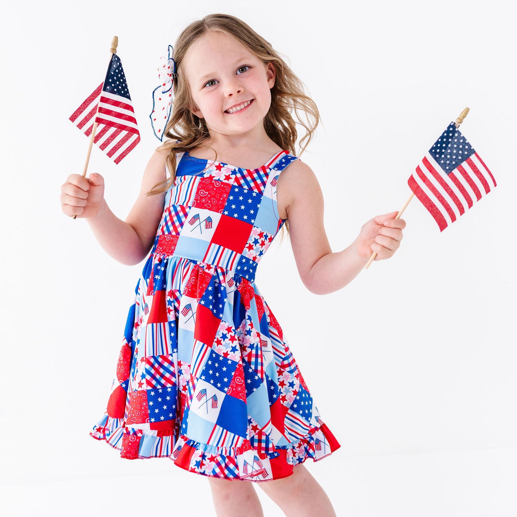 Young girl in a red, white, and blue patchwork dress holding two small American flags on a white background