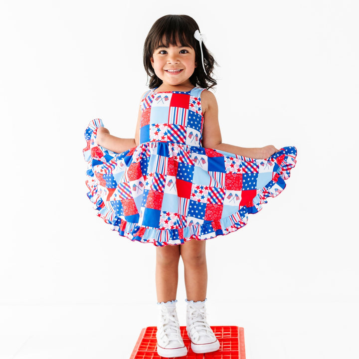 little girl posing in red, white and blue patchwork print sleeveless twirl dress with matching blue lettuce trim midi socks and white satin hair bow for the 4th of July