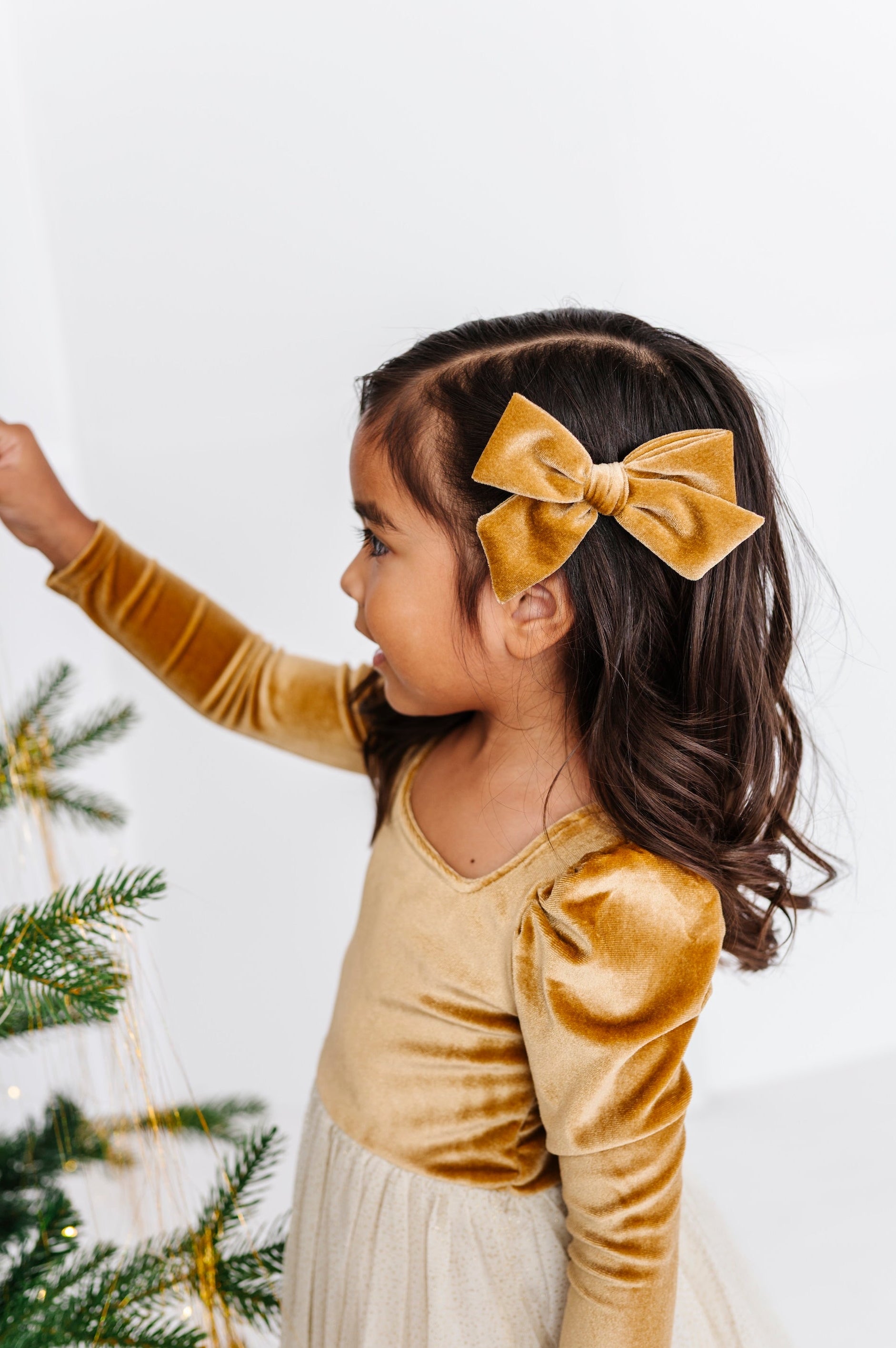 A young girl in a gold velvet long-sleeve dress and cream skirt is shown from the side, wearing a matching gold velvet bow headband. She is standing next to a decorated Christmas tree against a plain white wall.