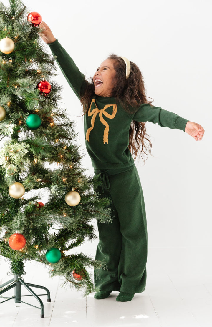 little girl decorating Christmas tree in forest green sweater pant set with gold bow detail on pullover