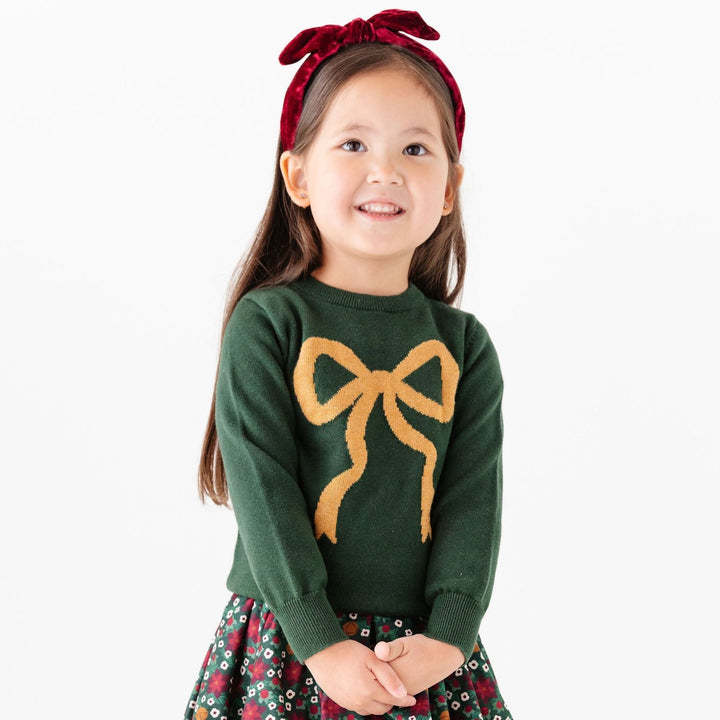 little girl wearing forest green sweater with gold bow and christmas poinsettia dress with matching red tights and headband