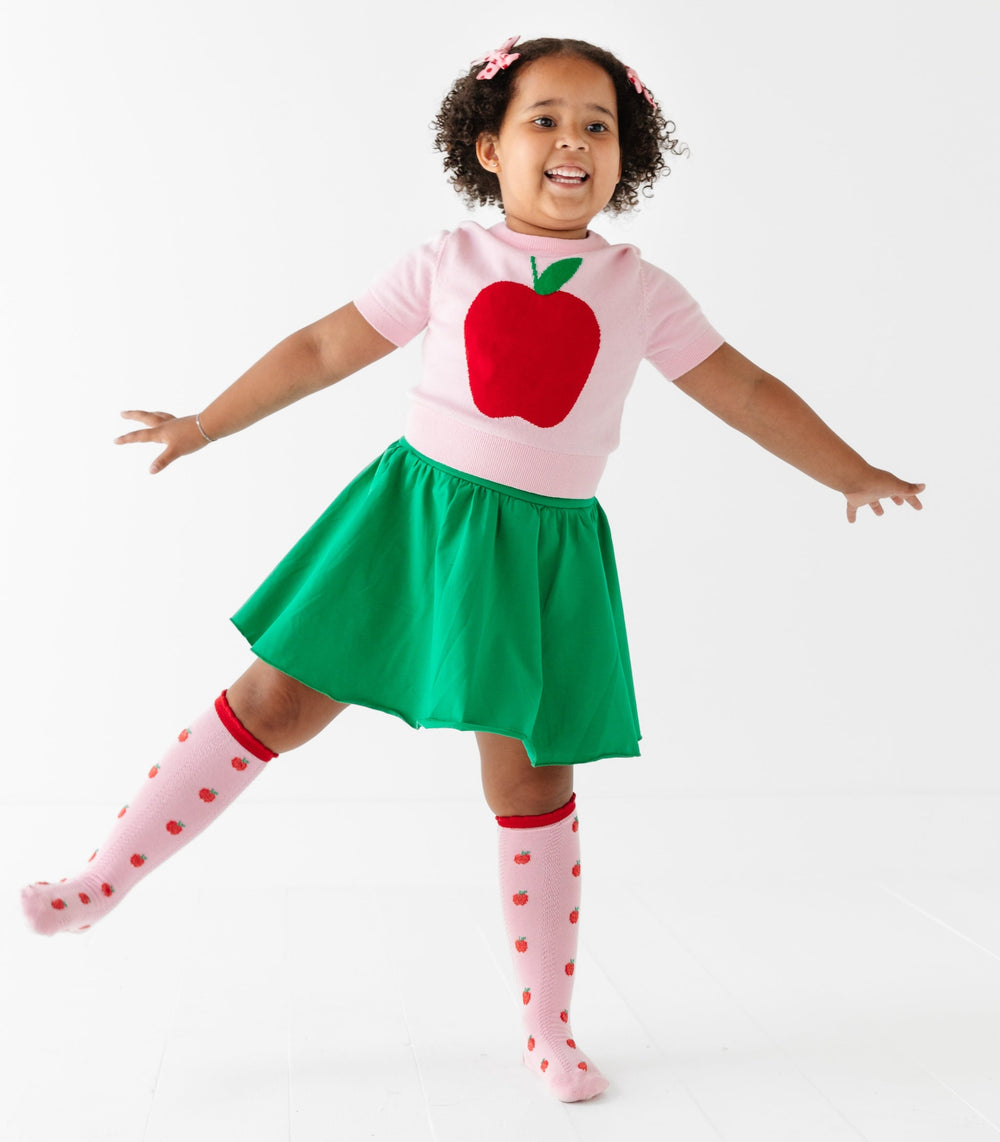 girl in back to school outfit with green cotton skort, pink short sleeve sweater with apple on it and matching pink knee highs with red apples on them