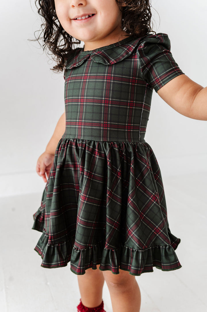 A toddler girl wearing a dark green plaid dress with red and white stripes, featuring a ruffled collar, short sleeves, and a tiered ruffled skirt.