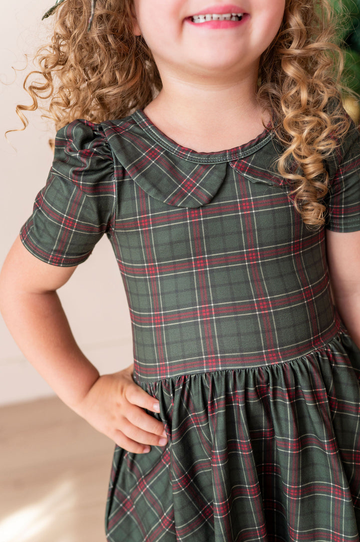 A close-up of a young girl wearing a dark green plaid dress with red and white stripes, featuring short puff sleeves and a Peter Pan collar.